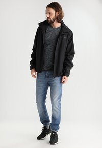 Veste imperméable noire avec capuche, t-shirt gris à motif, jeans bleu clair et baskets noires. Tenue décontractée au design fonctionnel.