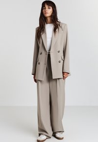 Femme se tenant contre un fond blanc, portant un blazer beige à double boutonnage, un pantalon large assorti, une chemise blanche et des baskets blanches.