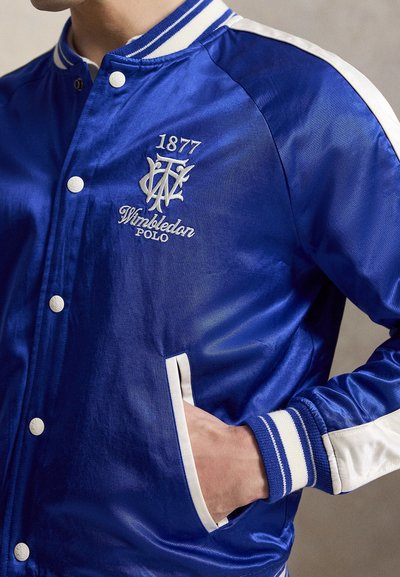 Μπλε σατέν varsity τζάκετ με λευκές ρίγες, κεντημένο «1877 Wimbledon Polo» στο στήθος και χέρι στην μπροστινή τσέπη.