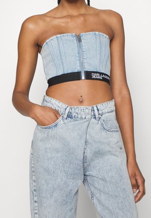 Kvinde iført lyseblå denim crop top med lynlås og stropper, samt højtaljede lyseblå denim jeans, med en synlig navlepiercing.