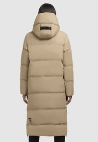 Lang beige pufferjakke med hætte, der har quiltede paneler, et logo-patch på ryggen og lynlåslommer i siderne. Glat tekstur.
