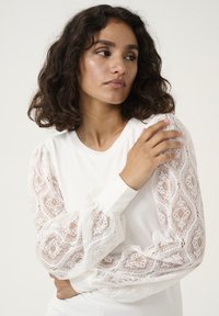 Long-sleeve top featuring a solid white body and sheer white lace sleeves with a floral pattern and gathered texture at the cuffs.