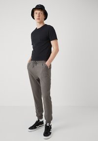Camiseta negra de manga corta, pantalones jogger grises con cintura elástica, y zapatillas deportivas negras de caña alta con suelas blancas. El modelo lleva un sombrero tipo bucket negro.