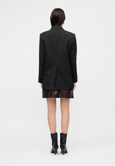 Μαύρο υπερμεγέθες σακάκι με μια μοναδική σχισμή στο πίσω μέρος, συνδυασμένο με καφέ δερμάτινη μίνι φούστα και λαμπερά μαύρα μποτάκια αστραγάλου.