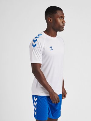 Man die naar rechts kijkt, draagt een wit sportshirt met korte mouwen met blauwe chevrons op de schouders en blauwe sportshorts.