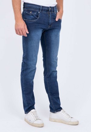 Homme portant un jean slim bleu foncé et des baskets blanches, debout devant un fond clair uni.