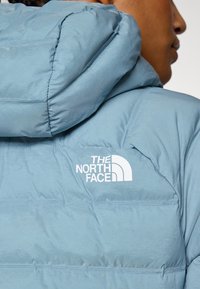 Light blue puffer jacket with a structured hood. Features include a smooth texture and a white logo on the left shoulder.