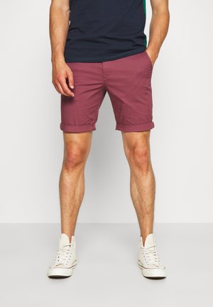 Bordeauxfarbene Baumwollshorts mit aufgerollten Bündchen, flachem Frontdesign und Seitentaschen, kombiniert mit weißen High-Top-Sneakern und einem dunkelblauen Hemd.