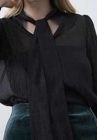 Blusa nera trasparente con costine verticali, design con fiocco al collo e dettaglio a taglio sul collo, abbinata a una gonna verde texturizzata.