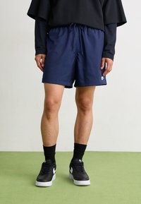 Navy blue swimming shorts with an elastic waistband and drawstring. Paired with black sneakers and black ankle socks on a green surface.