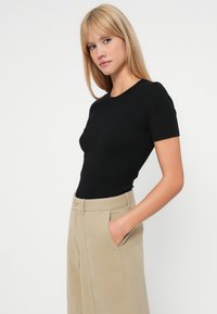 Top preto de manga curta em ribana combinado com calças bejes de cintura alta com bolsos frontais e fecho de botão. Fundo liso.