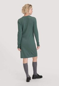 Grünes Strickkleid mit langen Ärmeln, Rundhalsausschnitt und einer Länge bis zur Mitte des Oberschenkels. Getragen mit grauen knielangen Socken und schwarzen Schuhen.
