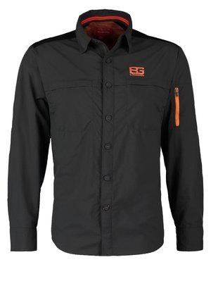 Mand går iført en mørkeoliven jakke med orange logo, grøn skjorte, brune bukser og sorte sko med gule snørebånd på hvid baggrund.