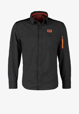 Mies kävelee päällään tumman oliivinvihreä takki, jossa on oranssi logo, vihreä paita, ruskeat housut ja mustat kengät, joissa keltaiset nauhat valkoisella taustalla.