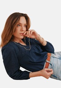 Marineblaue Bluse mit strukturiertem Stickmuster und Puffärmeln, kombiniert mit hoch taillierten Jeans und einem braunen Gürtel. Eine goldene Halskette hängt.