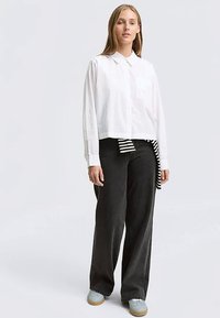 Camicia bianca con colletto, dal design corto, abbinata a pantaloni neri a gamba larga e vita alta, e scarpe azzurro chiaro. Dettaglio a righe visibile.