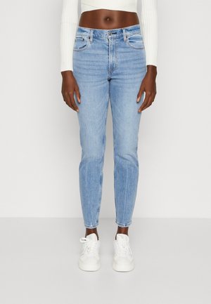 Forfra visning af person iført lyseblå højtaljede jeans med rå kanter, hvide sneakers og en kort hvid langærmet top.