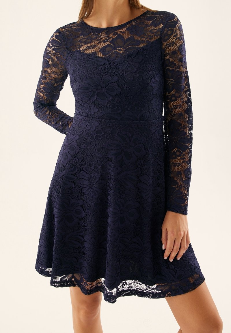 Femme portant une robe en dentelle florale bleu marine avec de longues manches transparentes et une taille cintrée, se tenant devant un fond uni.