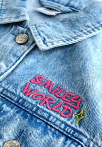Jachetă din denim cu nasture metalic și text brodat roz „SMILES WORLD”, alături de un mic detaliu cu stea galbenă.