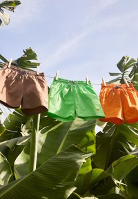 Tre paia di pantaloni corti pendono da mollette, in colori marrone, verde e arancione. Realizzati in tessuto leggero con fasce elastiche in vita.
