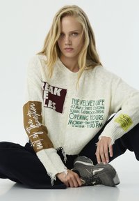 Pull blanc coupé avec tricot texturé ; présente des patchs imprimés en bourgogne et vert, et une manche marron avec un texte. Pantalon noir et baskets grises.
