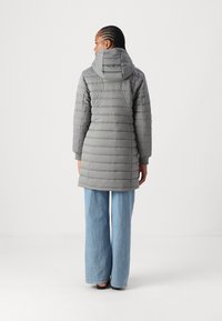 Grå polstret jakke med hette, med rett snitt og ribbestrikkede ermer. Brukt sammen med vide lyseblå jeans mot en ensfarget bakgrunn.