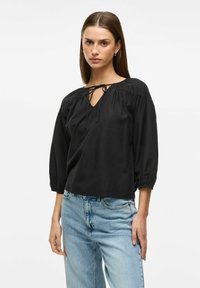 Melna blūze ar trīs ceturtdaļu piedurknēm, V-veida izgriezumu un saišu detaļu ap kaklu. Izgatavota no mīksta auduma ar brīvu piegriezumu un mazu tekstūru.