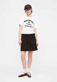 Mlada ženska, oblečena v belo majico z napisom "Weekend Max Mara", črno A-line krilo z žepi, temno kapo, bele nogavice in rjave čevlje Mary Jane.