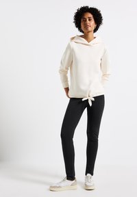 Ljust beige hoodie med knytbar midja och långa ärmar, kombinerad med svarta slim-fit-byxor och vita sneakers med beige detaljer.