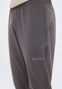 Κοντινό πλάνο ενός ατόμου που φοράει σκούρο γκρι παντελόνι Columbia με ελαστική μέση και πλαϊνές τσέπες, πάνω σε ουδέτερο φόντο.