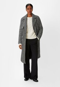 Manteau à motif pied-de-poule en noir et blanc, coupe oversize, avec des revers larges, poches sur le devant, porté sur un pull en laine torsadée crème et un pantalon noir.