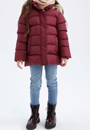 Enfant portant une doudoune bordeaux, un jean bleu clair, une écharpe beige et des bottes bordeaux brillantes à lacets, debout sur un fond blanc.