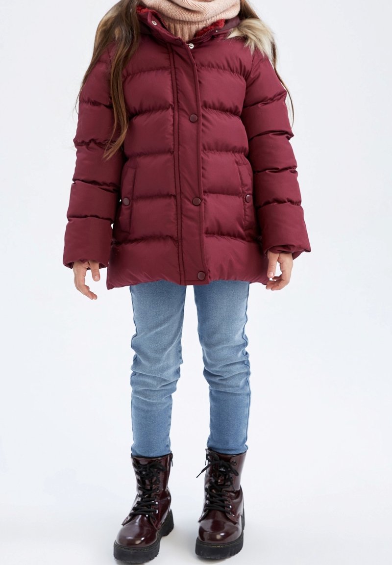 Enfant portant une doudoune bordeaux, un jean bleu clair, une écharpe beige et des bottes bordeaux brillantes à lacets, debout sur un fond blanc.