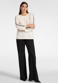 Vrouw in een crèmekleurige trui met grijze stikdetails op de mouwen, zwarte broek met wijde pijpen, staand met gekruiste armen tegen een witte achtergrond.