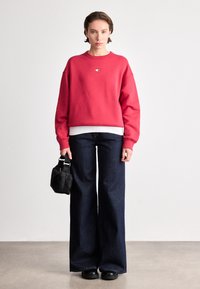 Sweatshirt rouge court, chemise blanche superposée en dessous, jean large noir, sac noir avec anses, et chaussures noires. Texture de tissu lisse.