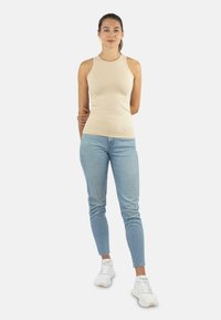 Beige ärmelloses Oberteil aus weichem Stoff, kombiniert mit hellblauen Skinny-Jeans und weißen Sneakers. Einfaches Design mit einer figurschmeichelnden Silhouette.