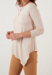 Top rosa chiaro a maniche lunghe con orlo curvo, realizzato in tessuto morbido, con maniche aderenti e una silhouette rilassata. Indossato con pantaloni beige.