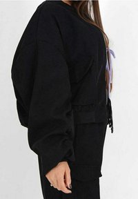 Vrouw in een oversized zwarte zip-up jas met grote zakken en lange mouwen, van opzij weergegeven tegen een effen achtergrond.