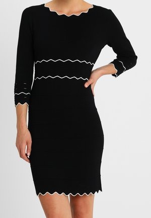 Black fitted dress with white zigzag trim on neckline, waist, cuffs, and hem, worn with one hand on hip and the other resting down.