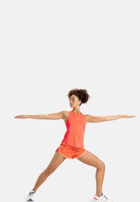 Sportliches Outfit mit einem leuchtend orangefarbenen Tanktop mit Logo und passenden orangenen Shorts mit weißen Akzenten, Modell in einer Kriegerpose.