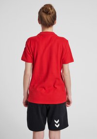 Rotes Baumwoll-T-Shirt mit kurzen Ärmeln und Kragen. Die Rückseite zeigt ein sauberes Design, kombiniert mit schwarzen Shorts, die ein weißes Zickzackmuster aufweisen.