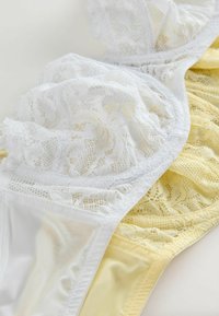 Hvide og lysegule blonde-bh'er, der overlapper hinanden og viser delikate blomsterdekorationer og gennemsigtige stofdetaljer på en ensfarvet baggrund.