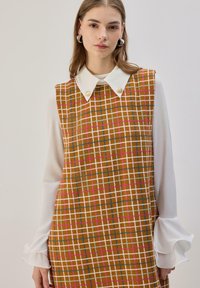 Robe sans manches à carreaux marron avec un col blanc orné de boutons dorés et des manches blanches douces à poignets volantés, présentant des motifs vibrants.