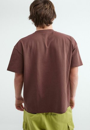 Persoon met kort bruin haar die een loszittend bruin t-shirt en lichtgroene cargoshorts draagt, van achteren gezien tegen een effen achtergrond.