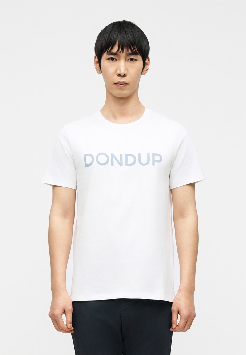 Άνδρας που φοράει λευκό κοντομάνικο μπλουζάκι μ ε τη λέξη "DONDUP" σε ανοιχτό μπλε γράμματα, στέκεται μπροστά σε ένα απλό λευκό φόντο.