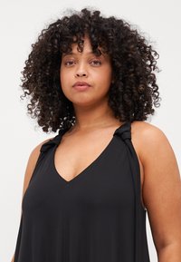 Rochie neagră fără mâneci, cu decolteu în V, având accente în formă de noduri pe bretelele umerilor și o textură de țesătură moale și fluidă.