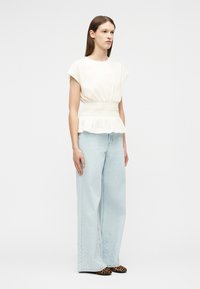 Mujer con una blusa blanca sin mangas con cintura ajustada, vaqueros anchos azul claro con patrones sutiles en el dobladillo y zapatos de estampado de leopardo.