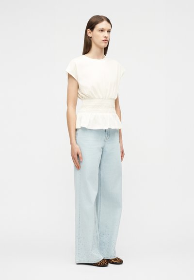 Γυναίκα που φοράει λευκό αμάνικο τοπ με σφιχτή μέση, ανοιχτό μπλε παντελόνι με φαρδιά πόδια και διακριτικά μοτίβα στο στρίφωμα, και παπούτσια με λεοπάρ σχέδιο.