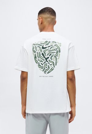 Mand iført hvid Nike Golf T-shirt med abstrakt grønt mønster og tekst "See you out there" på ryggen, stående mod en hvid baggrund.