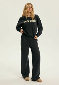 Czarna, oversize bluza z napisem "LAZY DAYS", w zestawie z luźnymi czarnymi spodniami. Miękki materiał, casualowy krój, do tego beżowe klapki. Minimalistyczne akcenty.